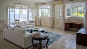 Un salón amplio con un sofá blanco, una mesa de centro de vidrio y un escritorio. Amplias ventanas ofrecen vistas al exterior.
