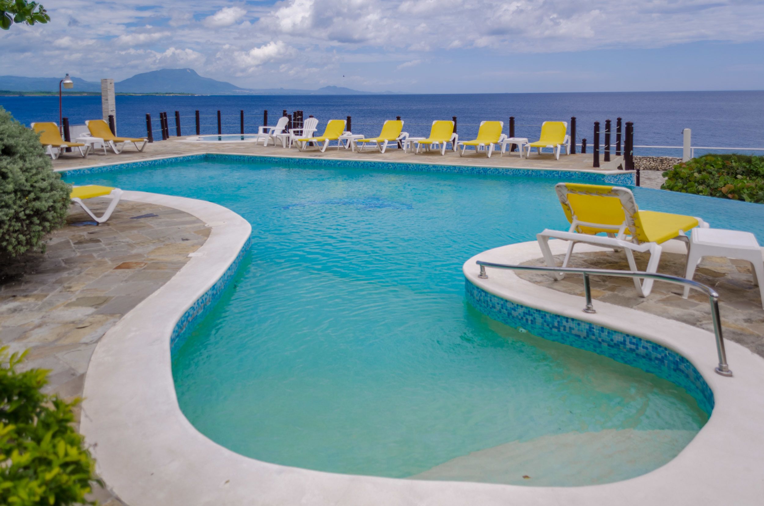 A swimming pool with yellow and white lounge chairs, surrounded by stone tiles and a view of the sea.