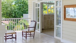 Un porche cubierto con una silla de madera y una mesita, una puerta blanca que da a un espacio cubierto, y vistas de follaje en el exterior.