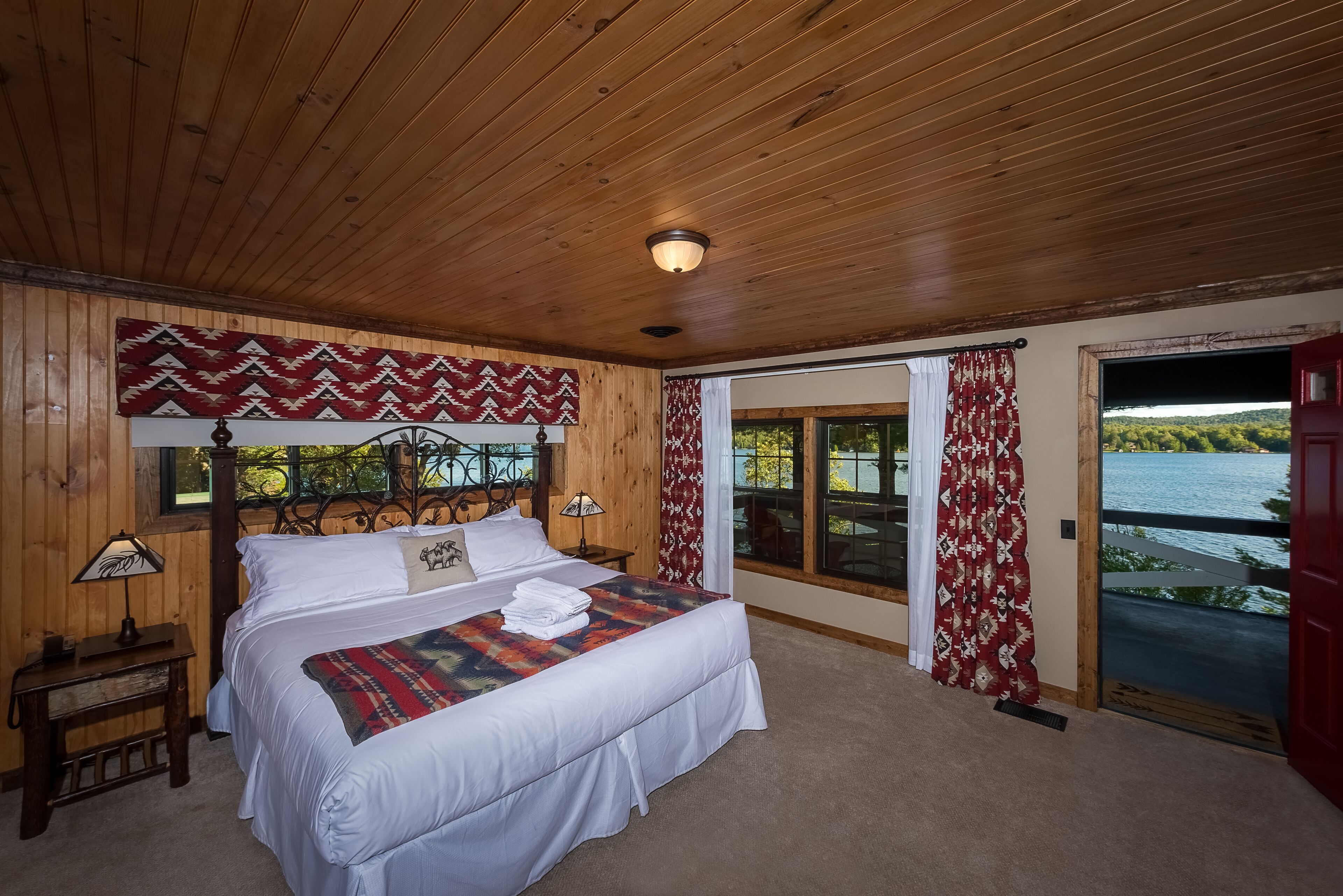 A wooden-paneled bedroom with a large bed, two bedside tables, a headboard, and a view of a lake through a sliding glass door.