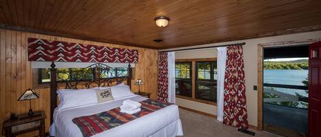 A wooden-paneled bedroom with a large bed, two bedside tables, a headboard, and a view of a lake through a sliding glass door.