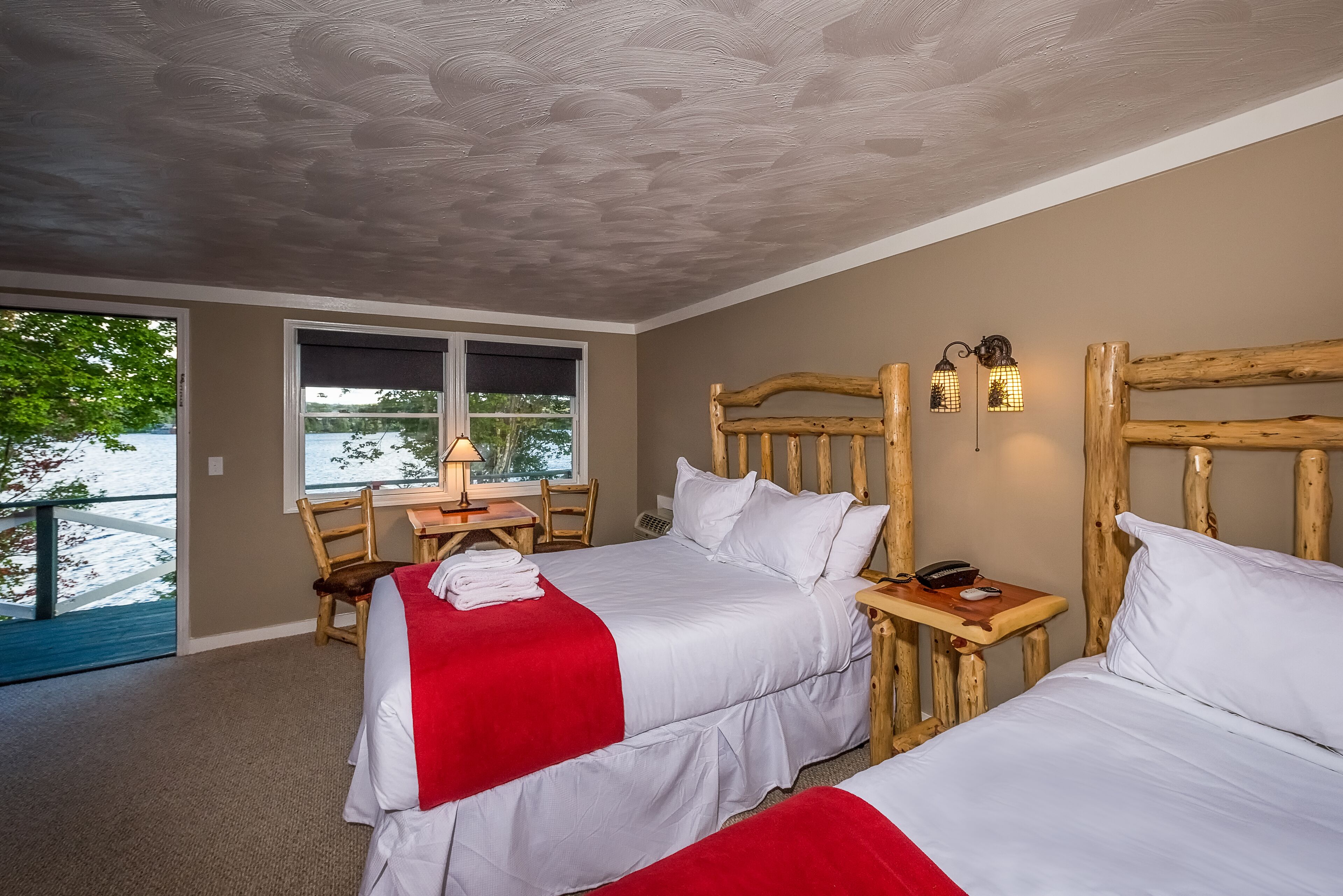 A hotel room with two beds, a wooden headboard, a desk with a chair, a lamp, and a view of the outdoors.