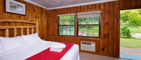 A wooden-paneled room with a bed, a framed picture, and a window with a view of a garden.