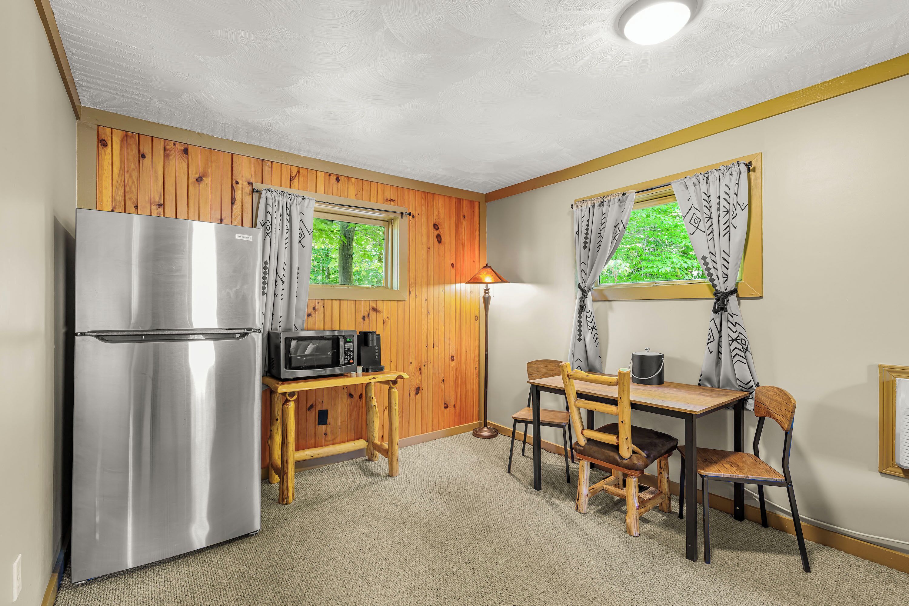 A compact kitchen with a stainless steel refrigerator, a microwave, a wooden table with chairs, and a window with patterned curtains.