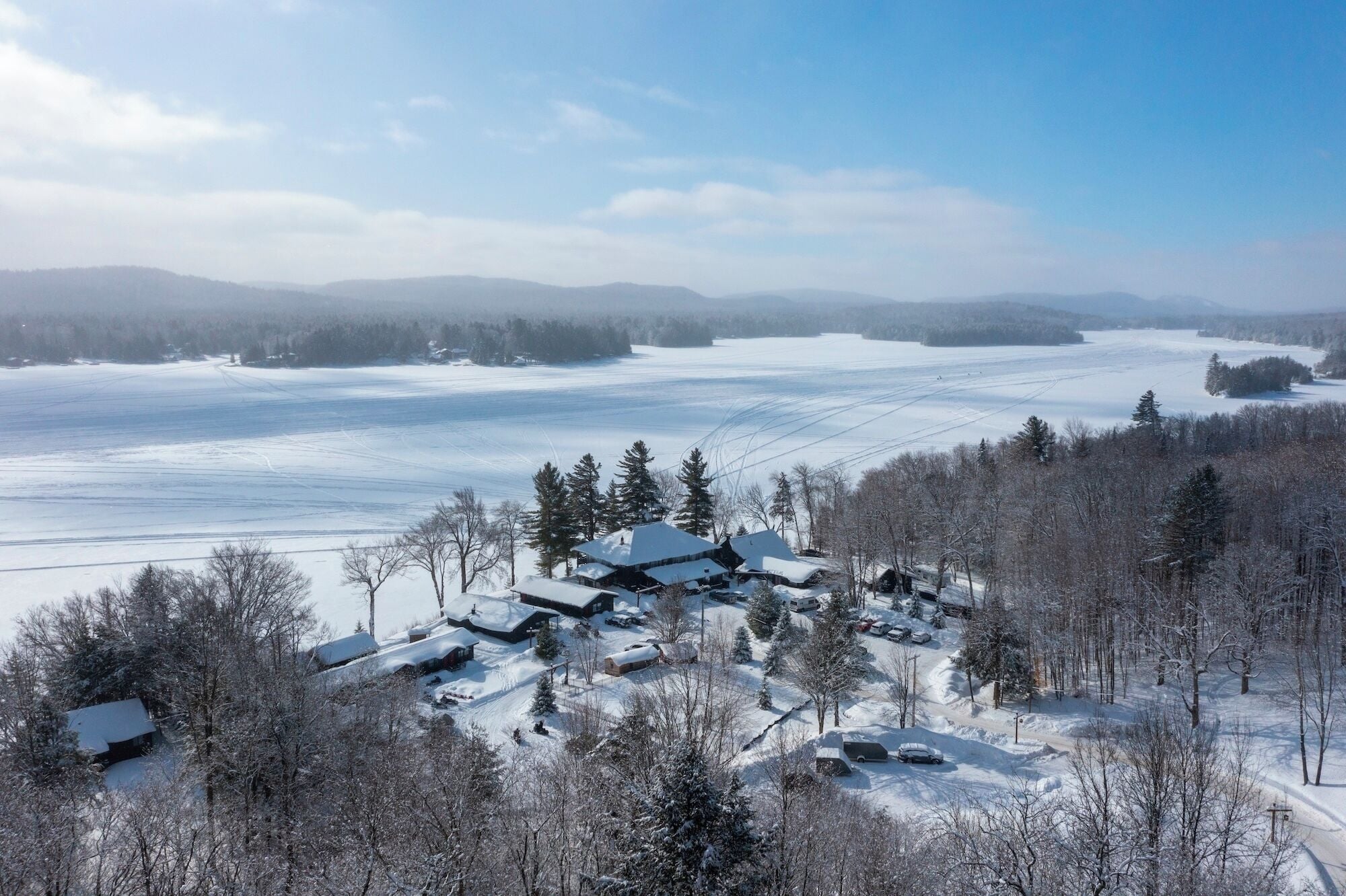 Een besneeerd landschap met een groep gebouwen, waarschijnlijk een resort of lodge, omringd door bomen en een bevroren meer.