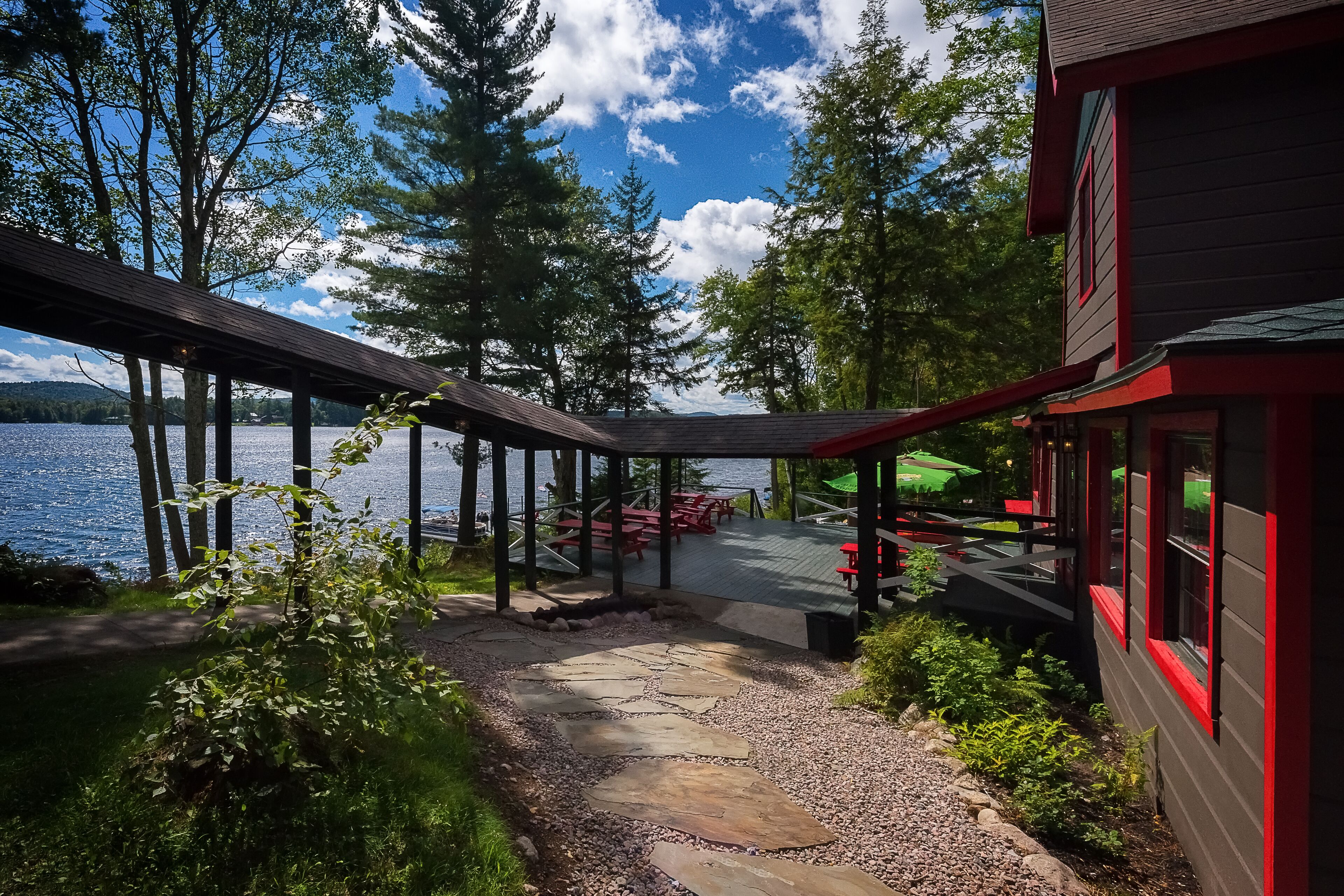 A lakeside deck with a stone pathway leading to outdoor seating, surrounded by trees and a building with red trim.