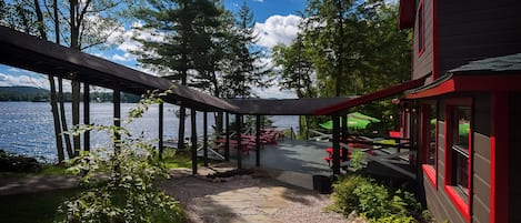 A lakeside deck with a stone pathway leading to outdoor seating, surrounded by trees and a building with red trim.