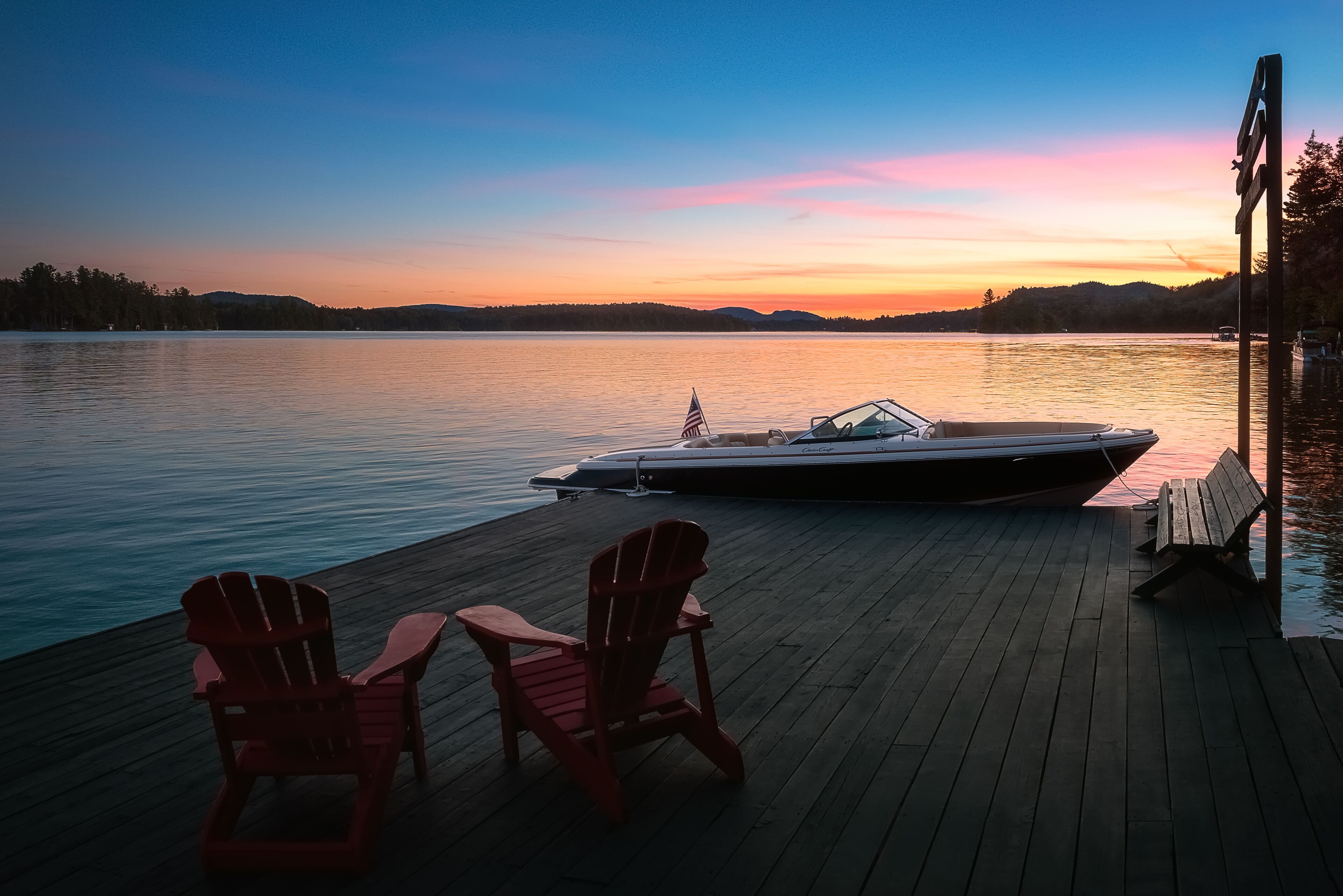 A wooden dock with two red Adirondack chairs and a boat docked at the end.