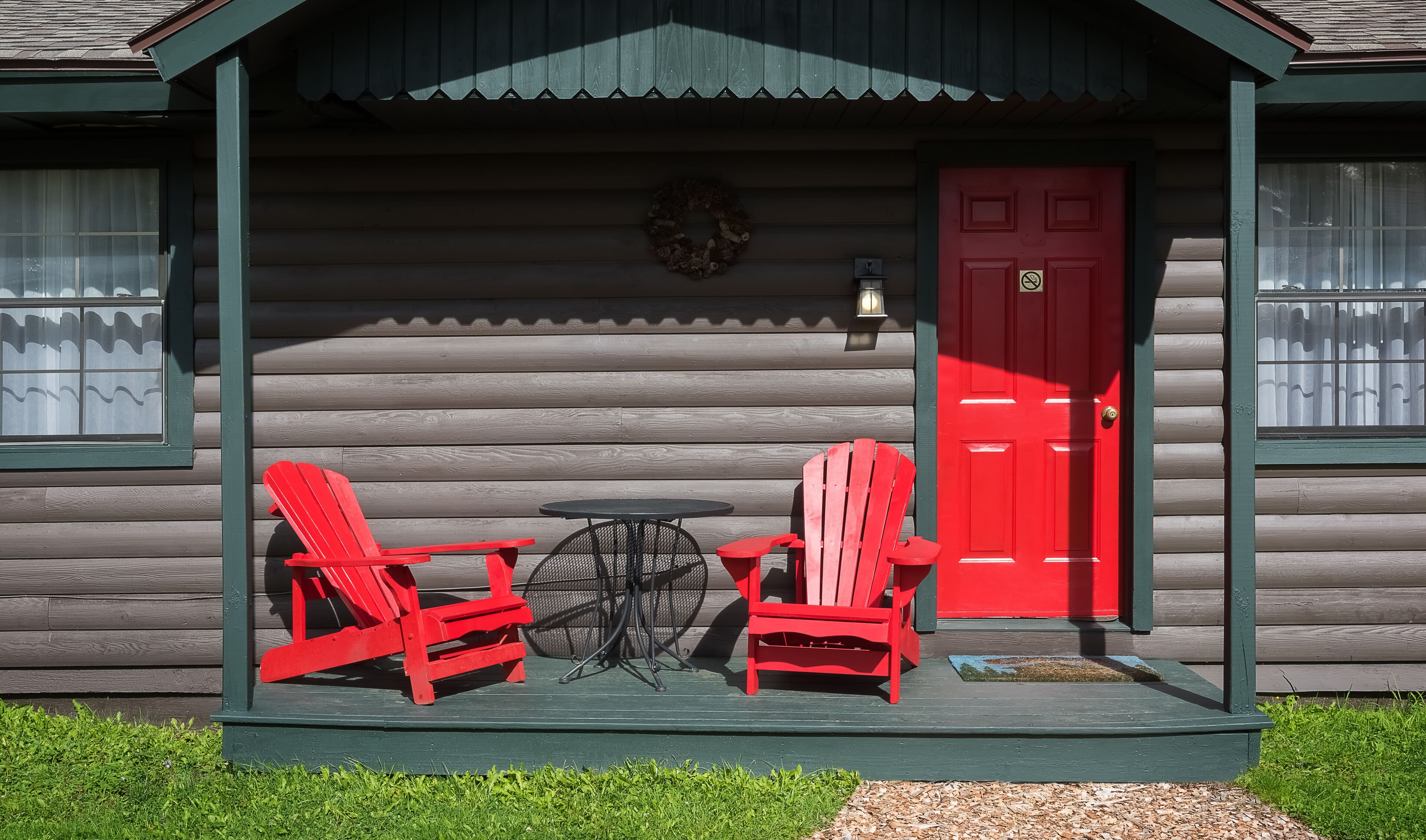 A log cabin with a porch, two red Adirondack chairs, a small round table, and a red door.