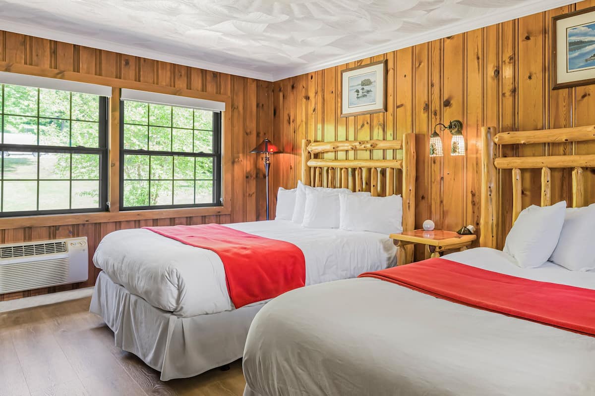 A room with two beds, wooden walls, large windows, and a ceiling fan.