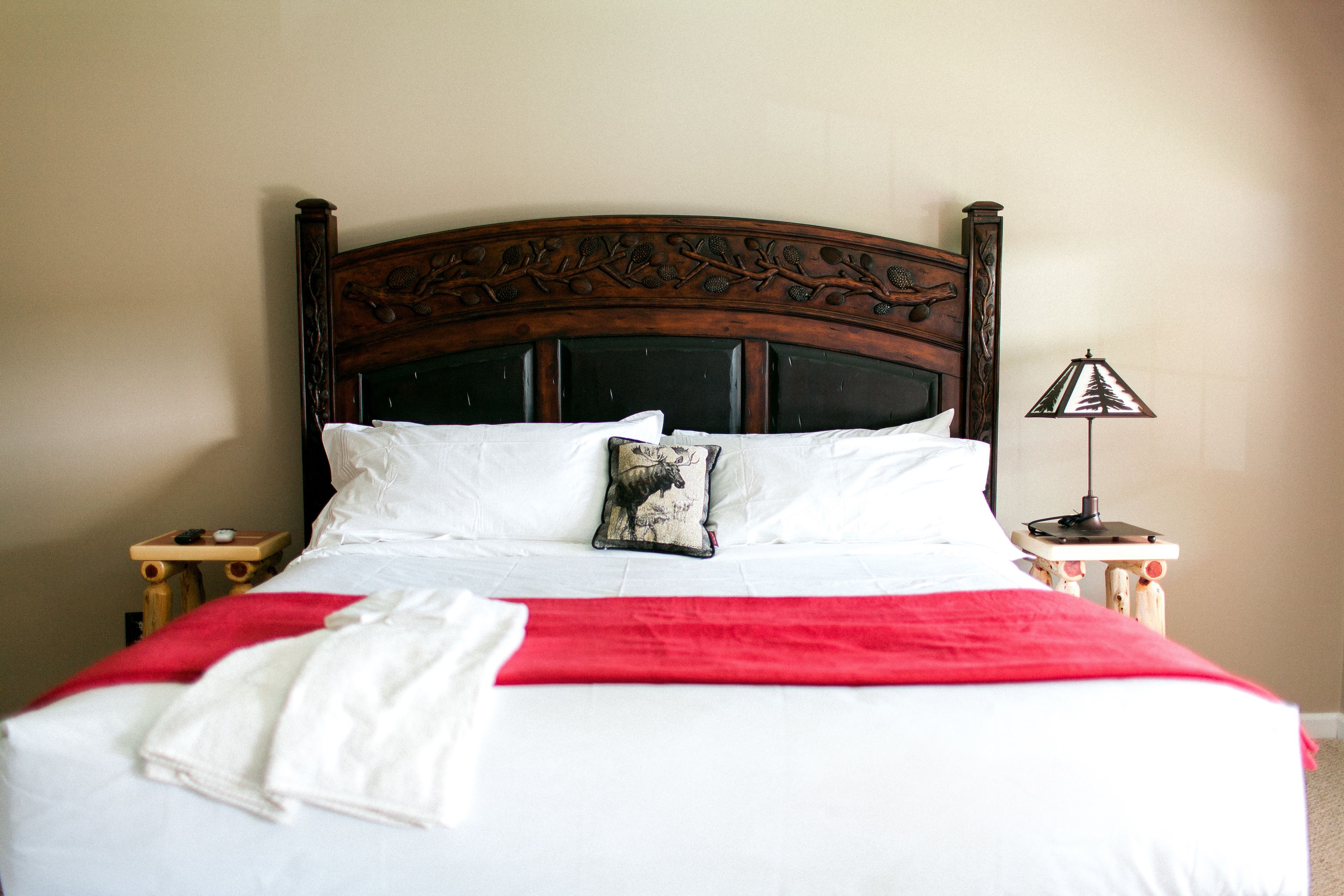 Una cama bien tendida con sábanas blancas y una manta roja, un cojín decorativo y una mesita de noche con una lámpara.