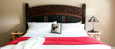 Una cama bien tendida con sábanas blancas y una manta roja, un cojín decorativo y una mesita de noche con una lámpara.