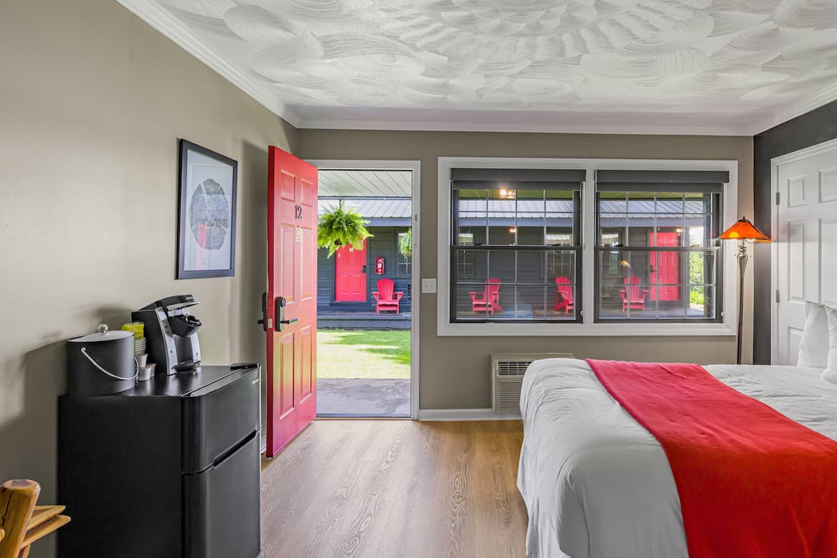 A hotel room with a bed, a desk with a coffee machine, a red door, and a window with a view of the outside.