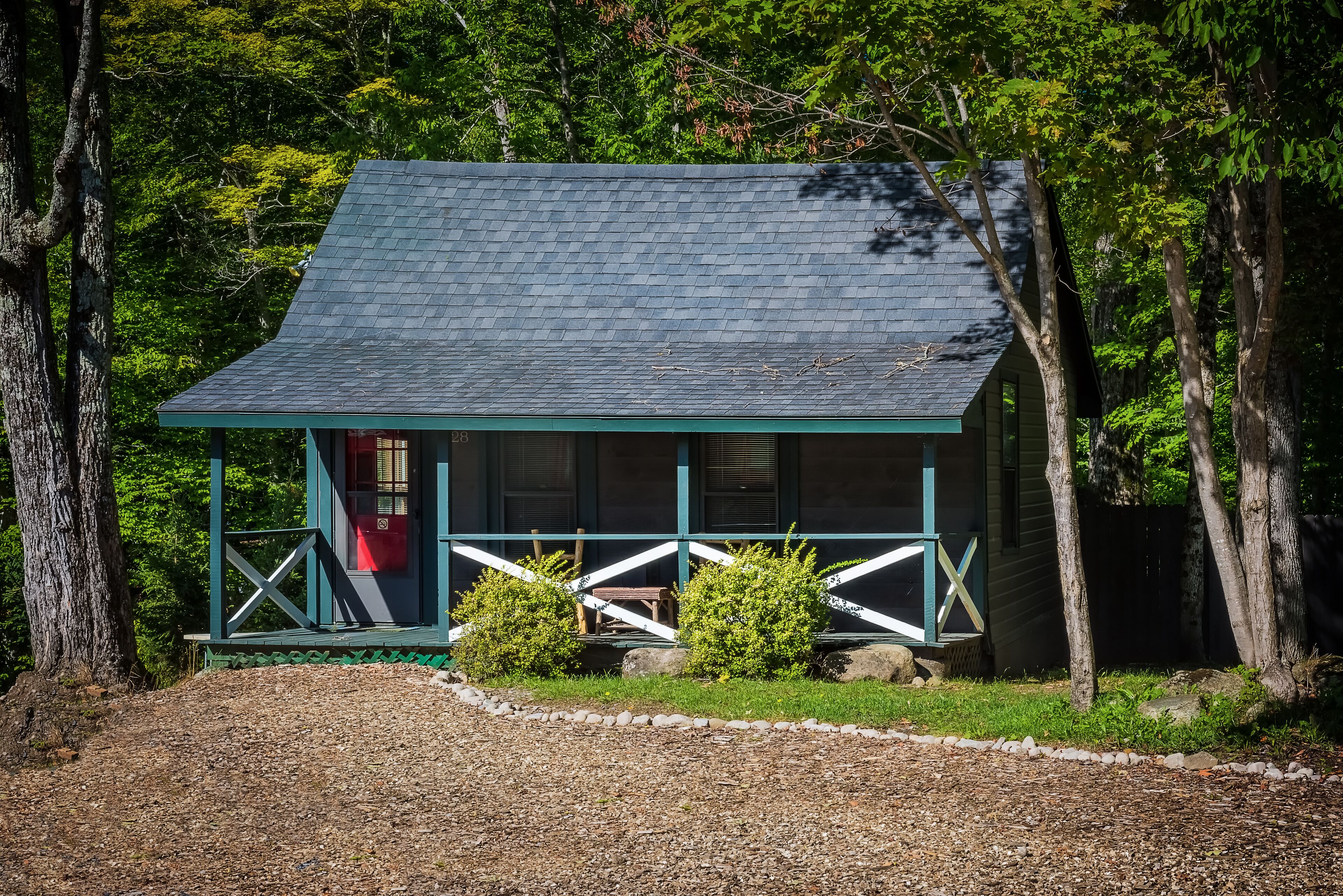 Een chalet met schuin dak, een veranda met een leuning en een kleine voortuin met een grindpad dat naar de ingang leidt.