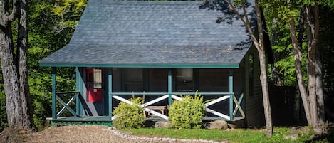 A cabin with a sloped roof, a porch with a railing, and a small front yard with a gravel path leading to the entrance.