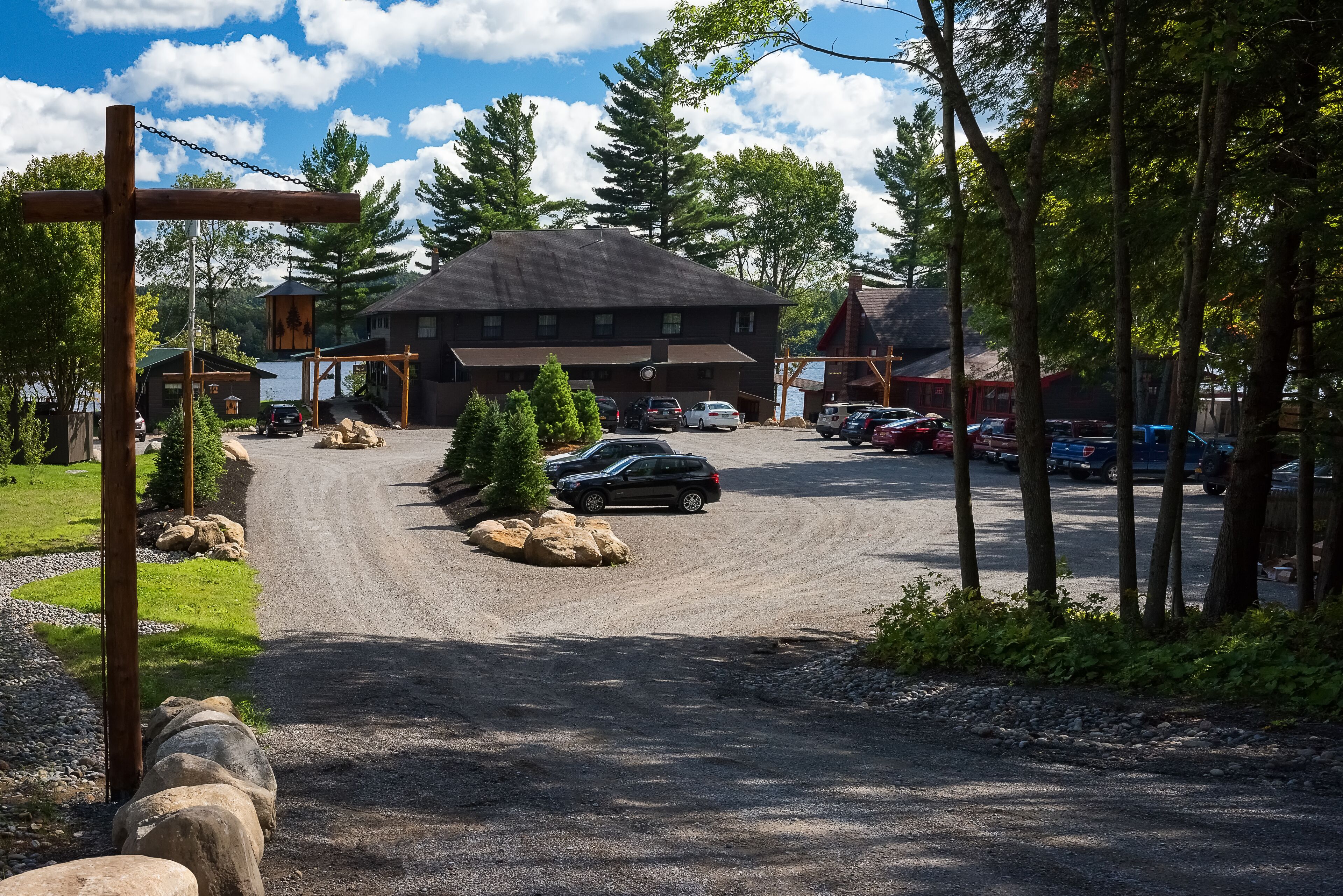 A gravel driveway leading to a building with a dark roof, surrounded by trees and parked cars.