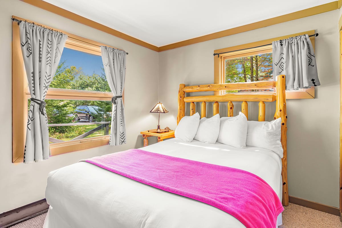 A bedroom with a wooden headboard, white bedding, a pink throw blanket, and a view of the outdoors through a window.