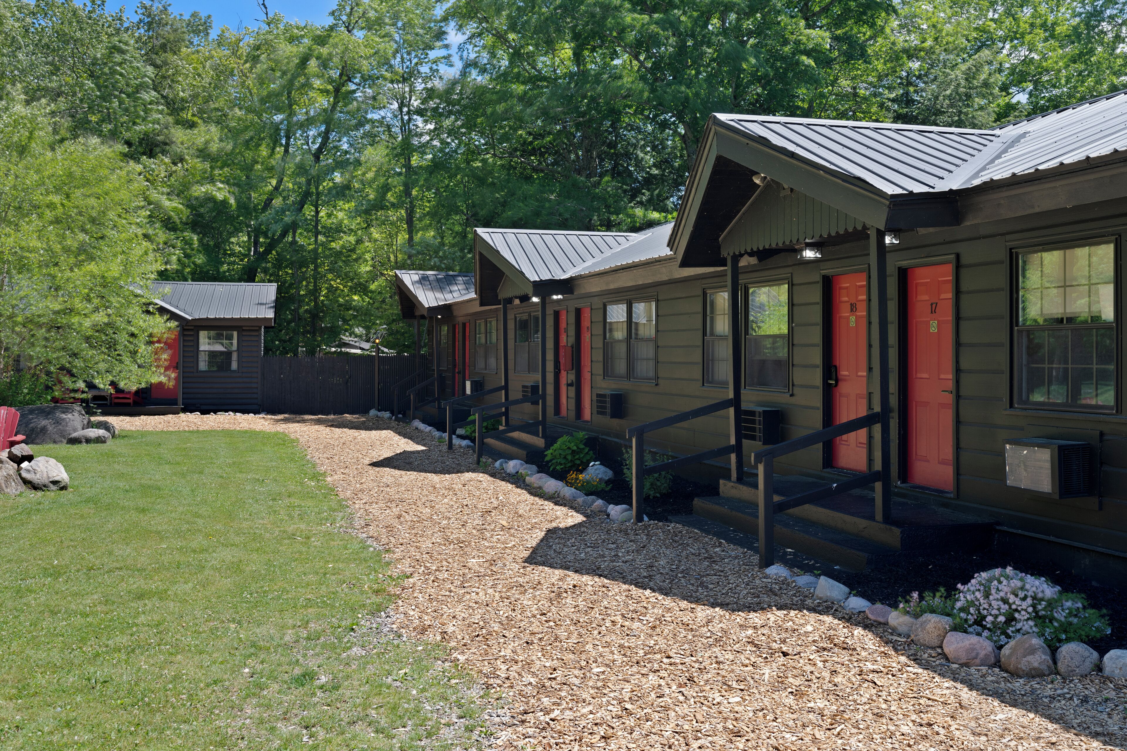 A row of cabins with metal roofs, wooden exteriors, and red doors, surrounded by a gravel path and greenery.