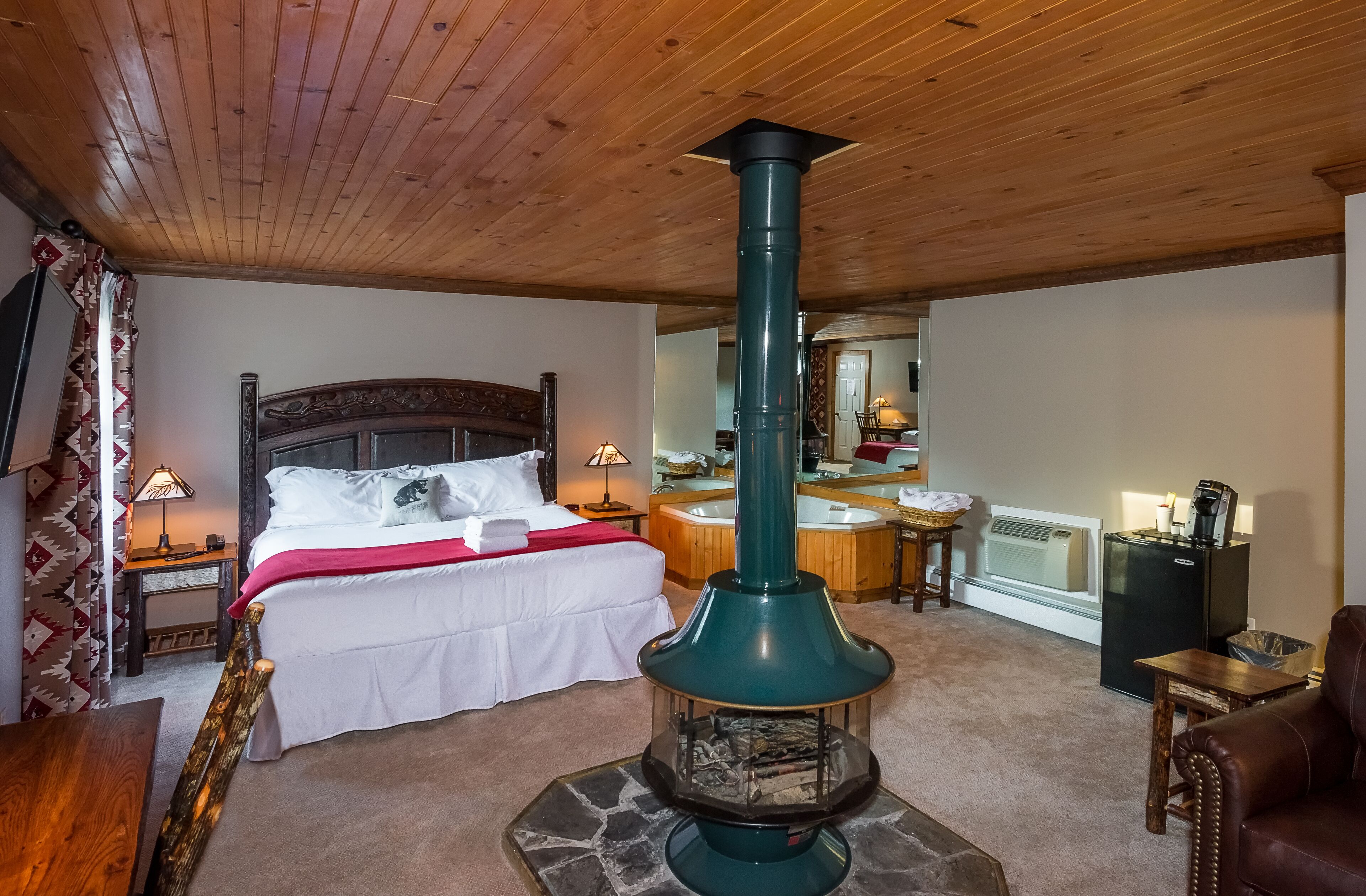 A cozy room with a bed, a wooden ceiling, a stone fireplace, a bathroom with a bathtub, and a refrigerator.