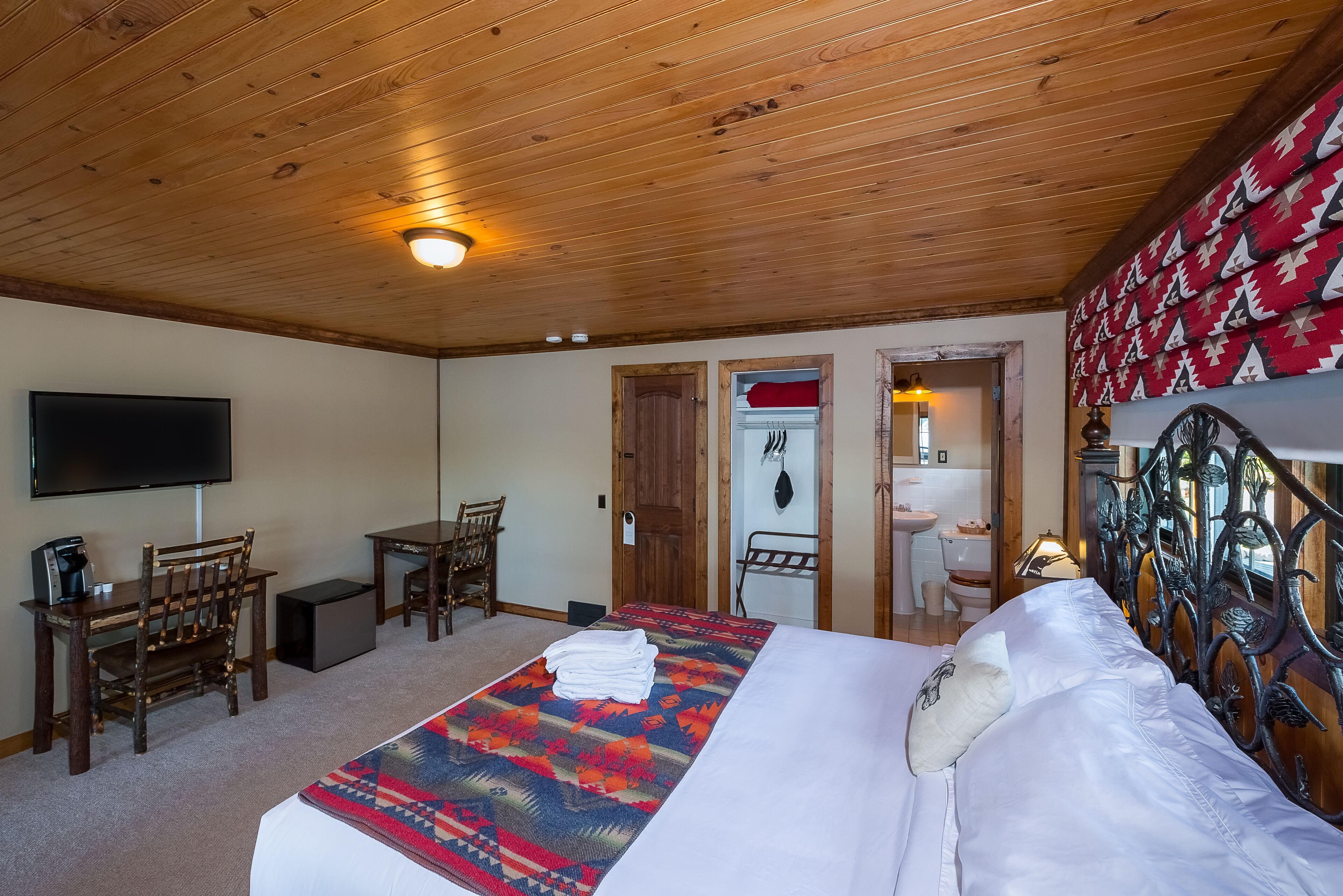 A hotel room with a wooden ceiling, a bed with a patterned blanket, a desk with a chair, a television, and a bathroom visible through an open door.
