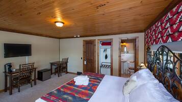 A hotel room with a wooden ceiling, a bed with a patterned blanket, a desk with a chair, a television, and a bathroom visible through an open door.