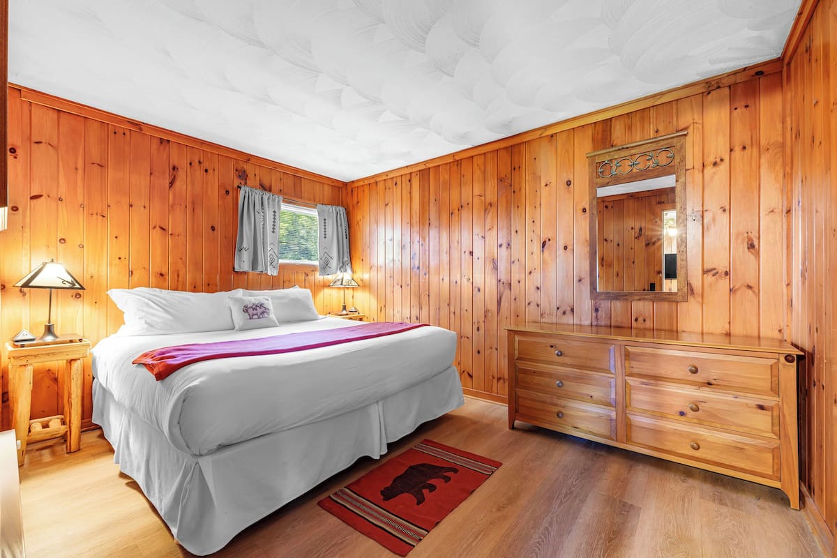 A bedroom with wooden walls, a bed with white bedding, a wooden dresser, and a red rug with a bear design.