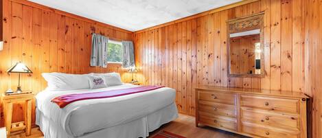 A bedroom with wooden walls, a bed with white bedding, a wooden dresser, and a red rug with a bear design.