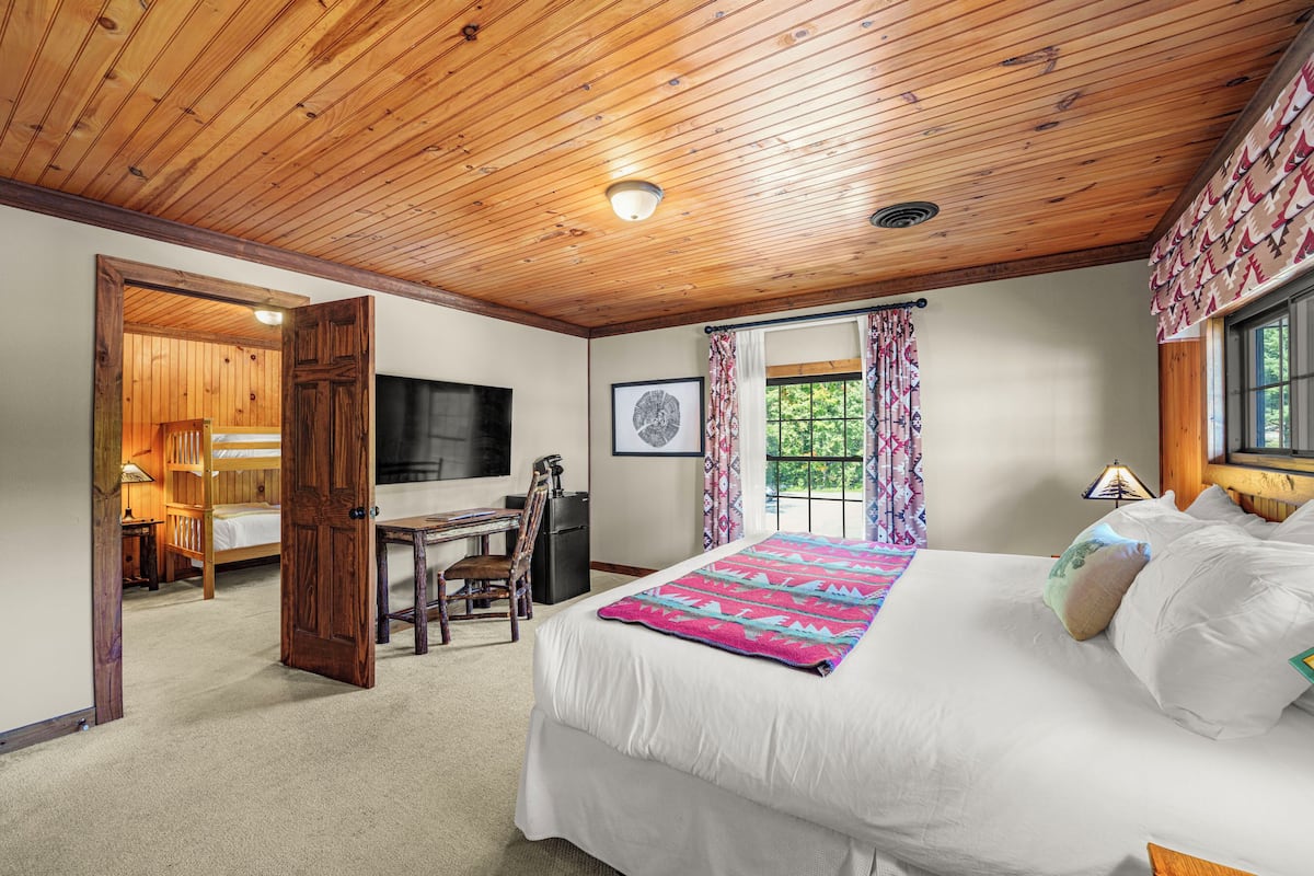 A bedroom with a wooden ceiling, a bed with white bedding, a desk with a chair, a television, and a window with curtains.