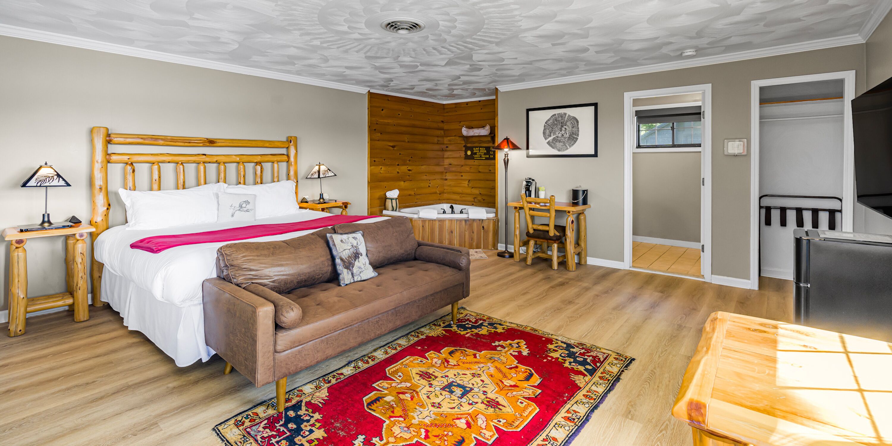 Une chambre d’hôtel comprenant un lit, une tête de lit en bois, un canapé en cuir marron, un tapis coloré, un bureau en bois, une chaise, un lavabo et un réfrigérateur.