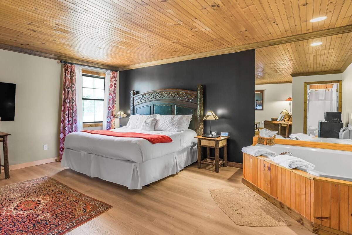 A spacious bedroom with a large bed, a wooden desk, a TV, and a bathroom with a bathtub and mirror.