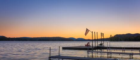 A calm lake at sunset with a dock and chairs.