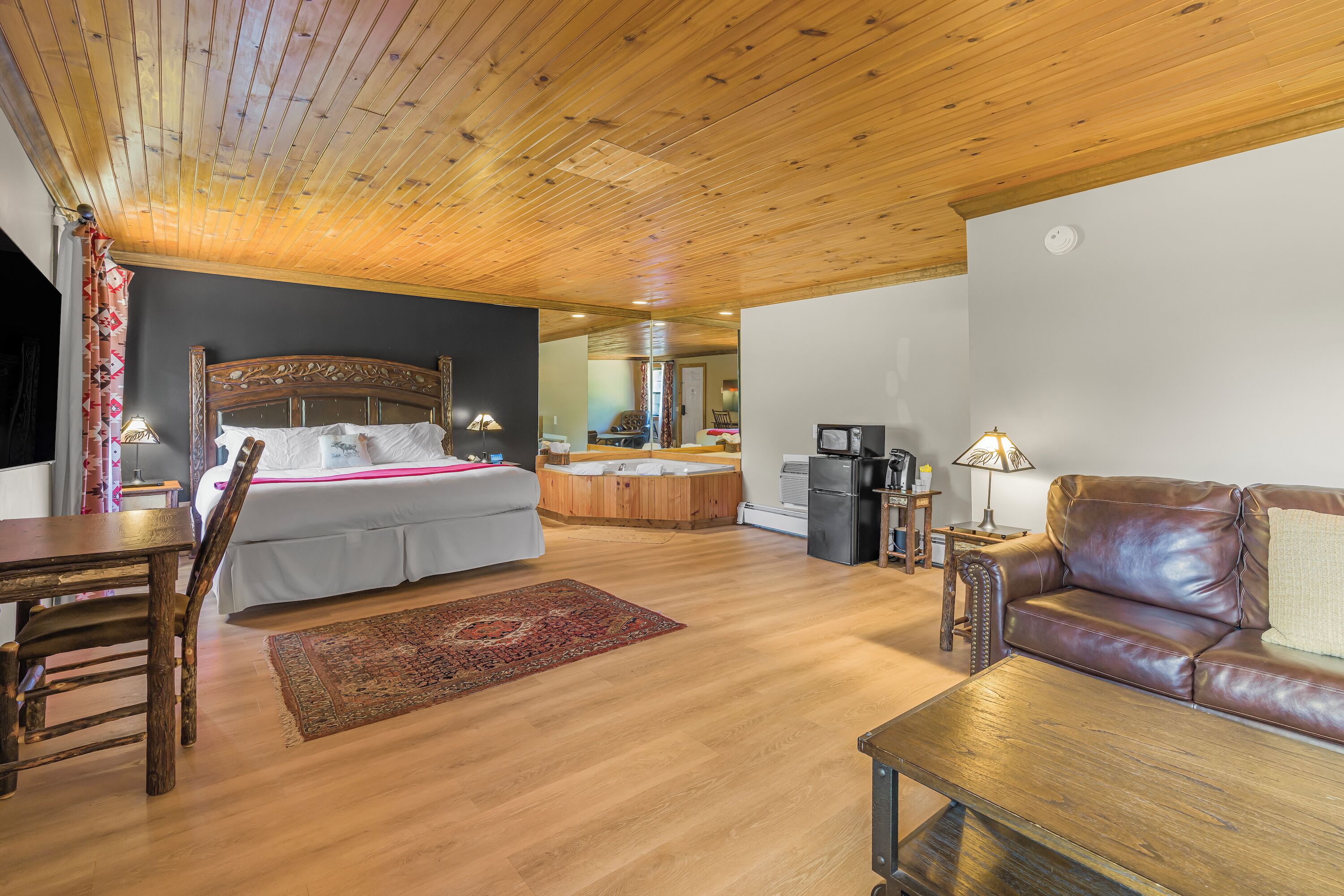 A room with a wooden ceiling, a bed with a headboard, a brown leather sofa, a wooden coffee table, and a small table with a lamp.