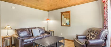 A living room with a wooden ceiling, a brown leather sofa, a matching armchair, a small table, and a floor lamp.
