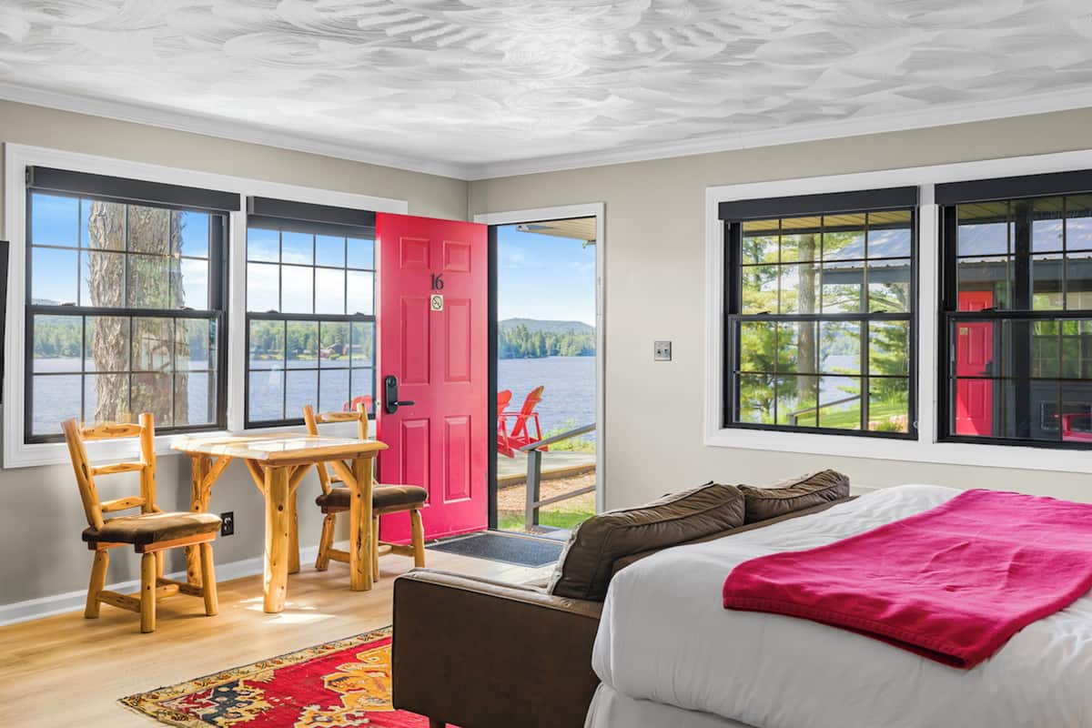 A room with a bed, a dining table with chairs, a red door, and large windows with a view of the outdoors.