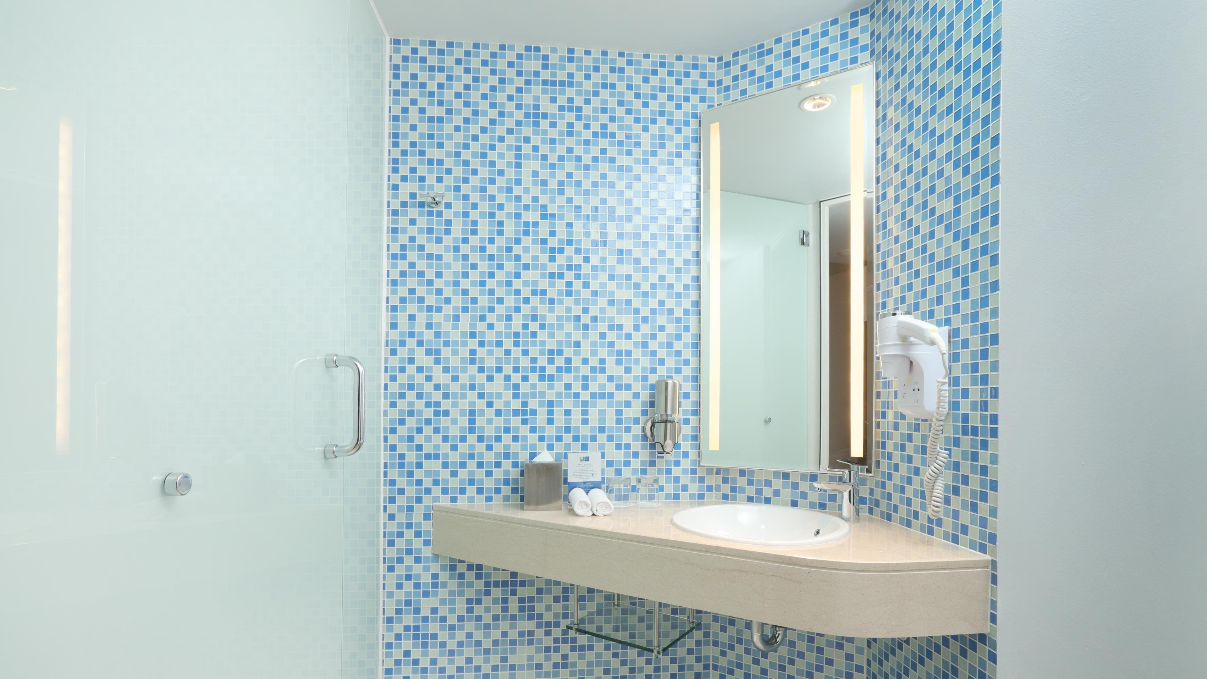 A bathroom with a blue mosaic tile wall, a glass shower enclosure, a white sink with a mirror, and a towel dispenser.