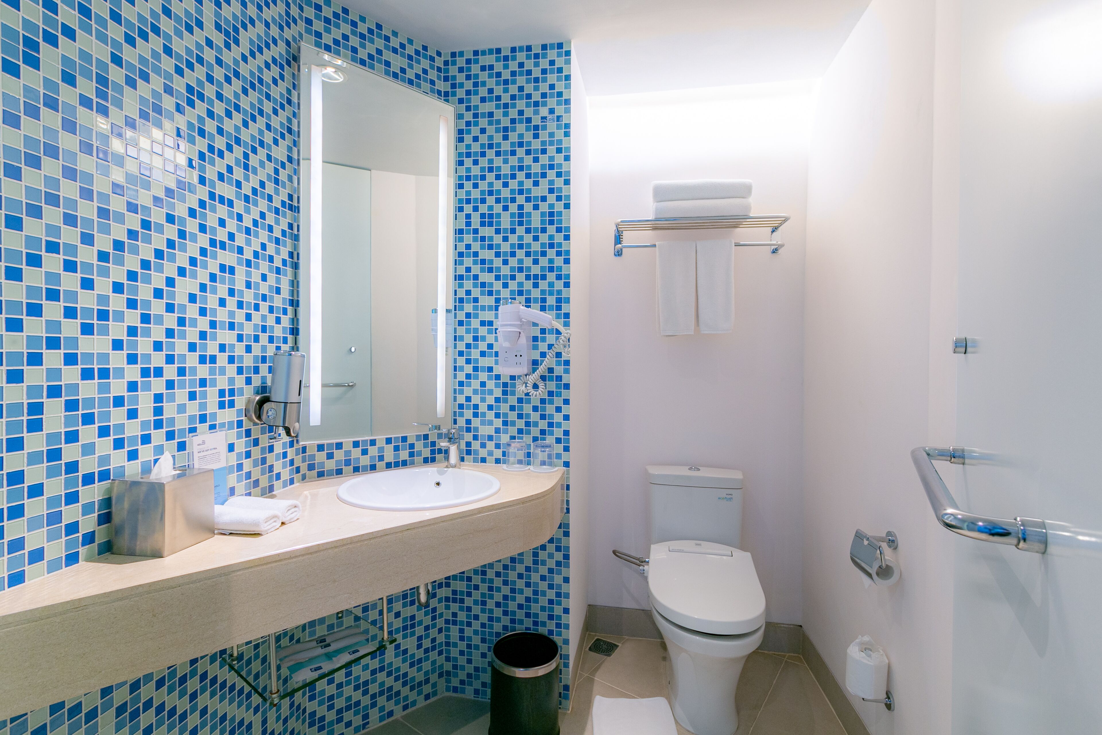 Een badkamer met een blauwe mozaïektegelwand, een witte wastafel, een spiegel, een toilet en een handgreep.