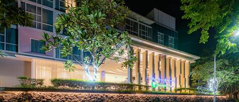 A modern hotel with a well-lit facade, a stone wall in front, and a large tree.
