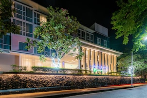 A modern hotel with a well-lit facade, a stone wall in front, and a large tree.
