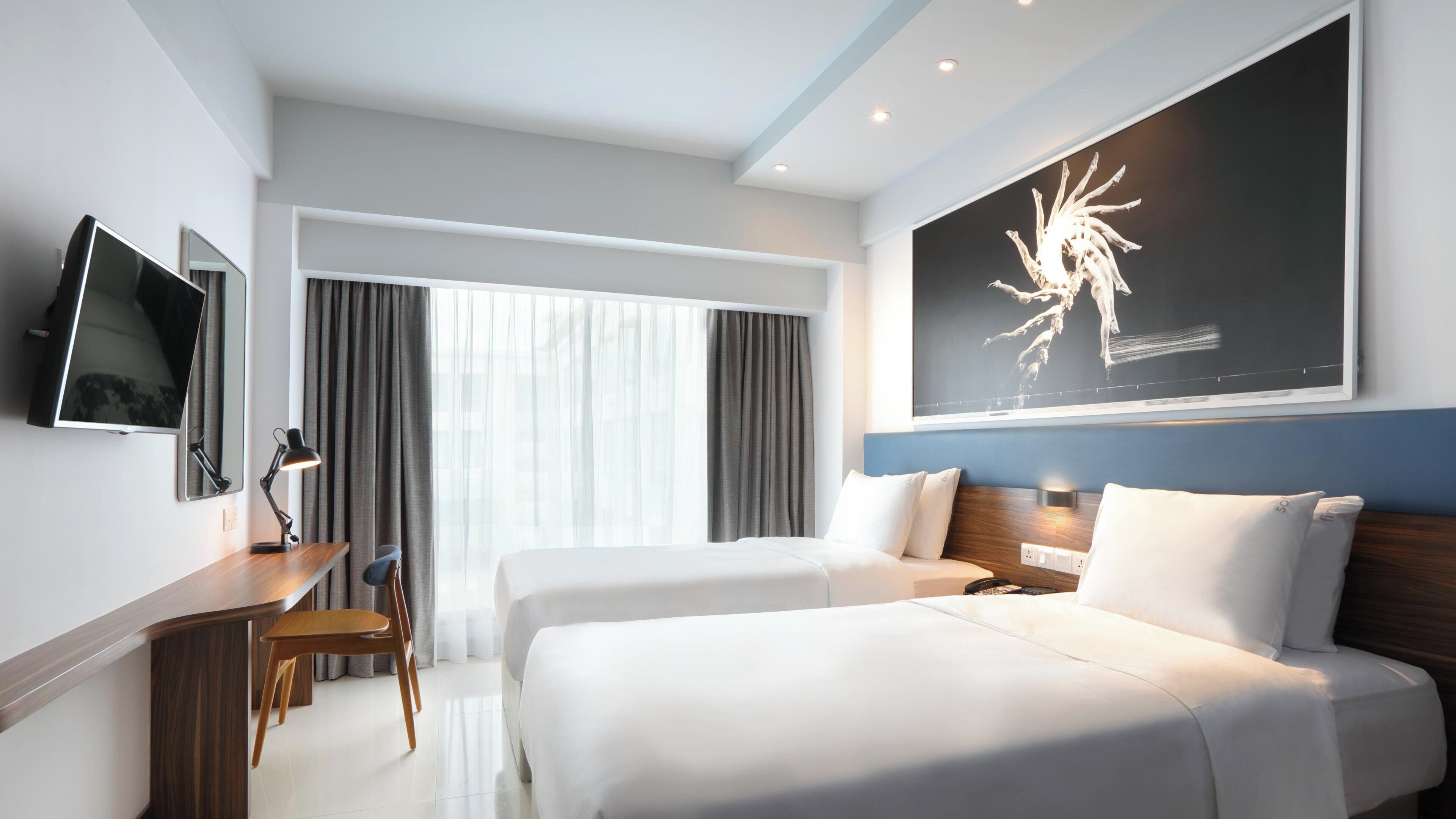 Habitación de hotel con dos camas, un escritorio con una silla, un televisor de pantalla plana y un cuadro abstracto grande en la pared.