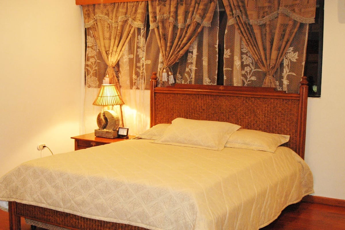 Cama bem arrumada com cabeceira, luminária de cabeceira e mesa de cabeceira com relógio.
