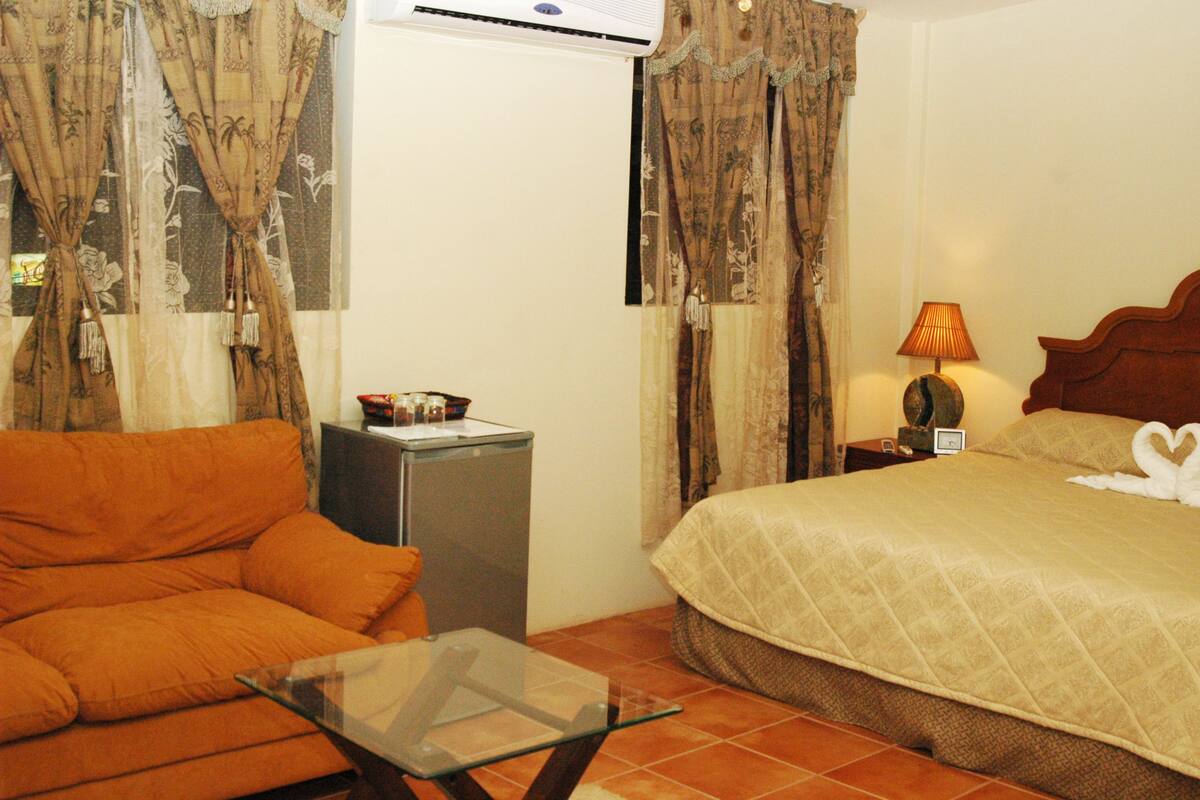 Quarto de hotel com uma cama, um sofá, uma mesinha, um abajur e cortinas.
