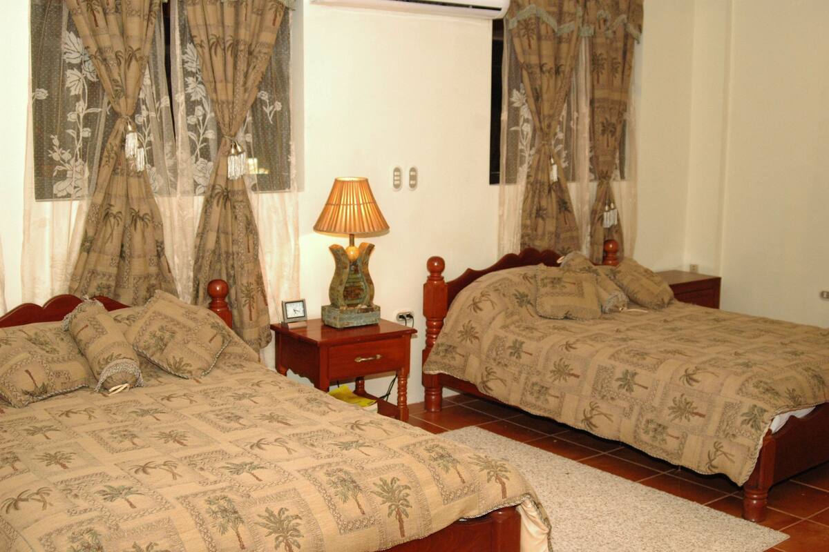 Quarto com duas camas de solteiro, mesa de cabeceira com abajur e cortinas com padrão floral.