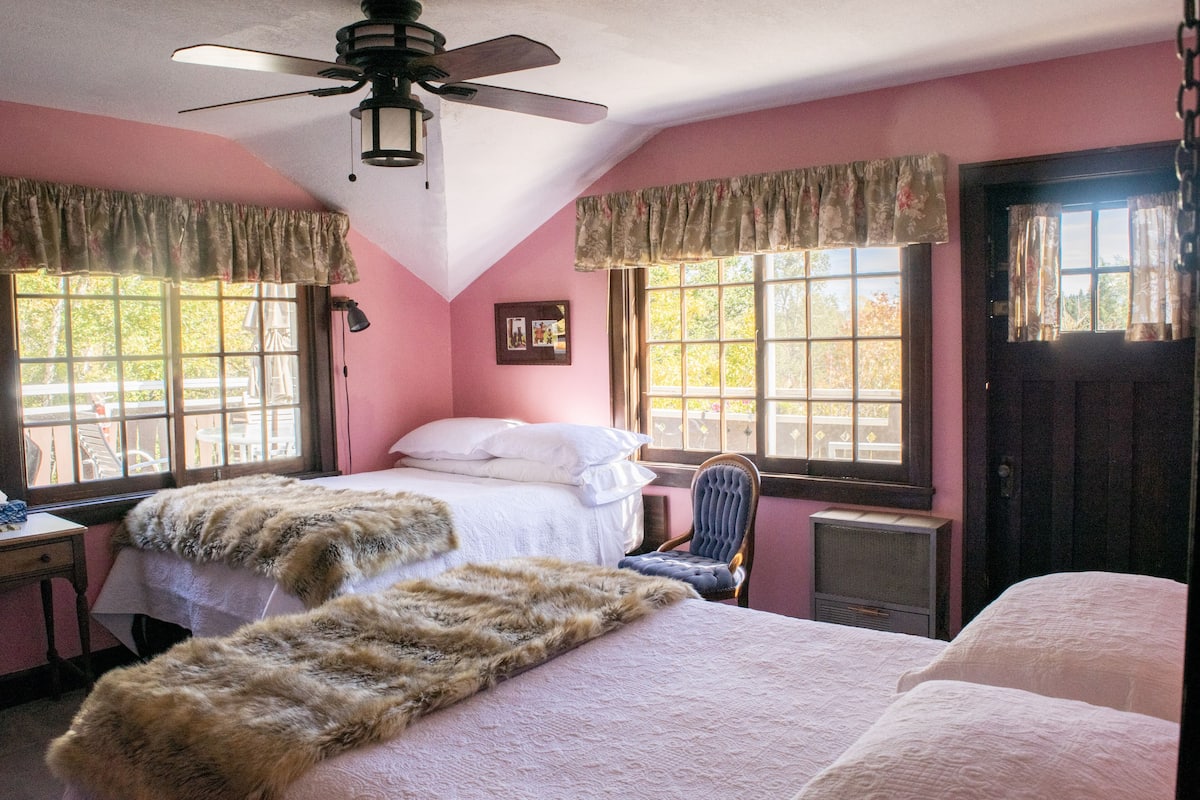 Une chambre avec deux lits, un ventilateur de plafond et de grandes fenêtres avec des rideaux.