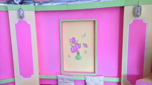 Une pièce aux couleurs vives, avec des tons rose et vert, un petit lit recouvert d’une couverture rouge, deux oreillers et une étagère fixée au mur, sur laquelle est posé un vase rempli de fleurs.