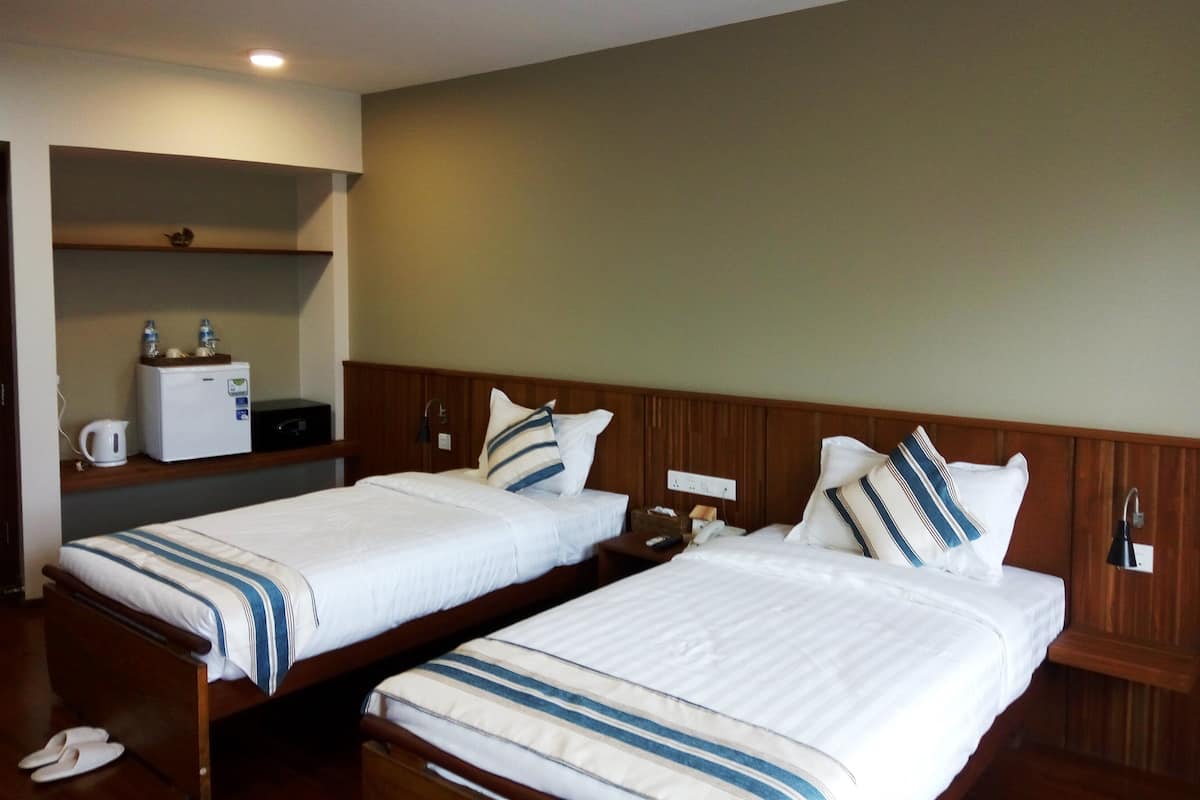 A hotel room with two single beds, a small kitchenette, a wooden headboard, a shelf with a kettle and water bottles, and a nightstand with a lamp.