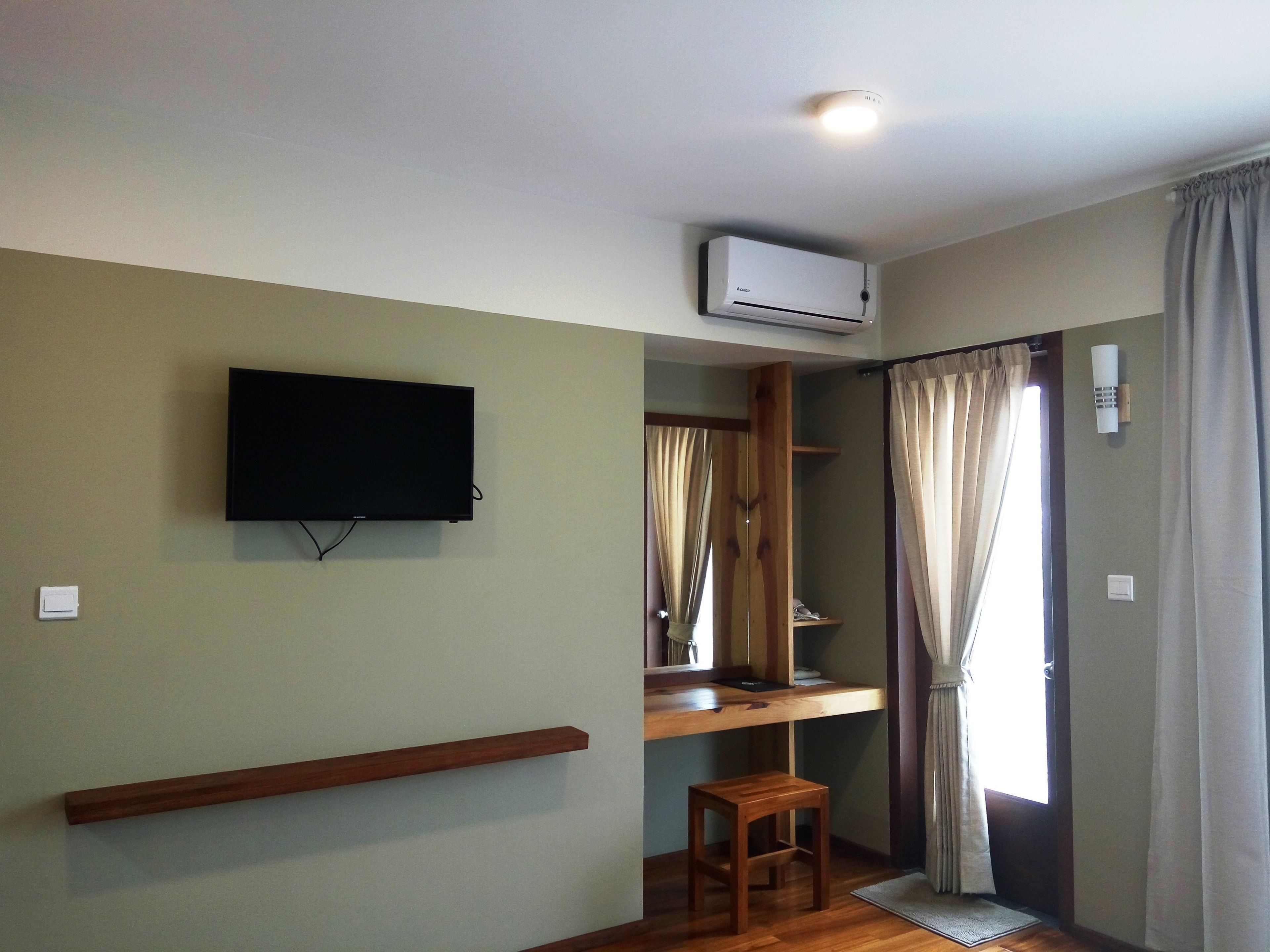 Una habitación con televisor de pared, aire acondicionado, un escritorio de madera, un taburete pequeño y una ventana con cortinas.
