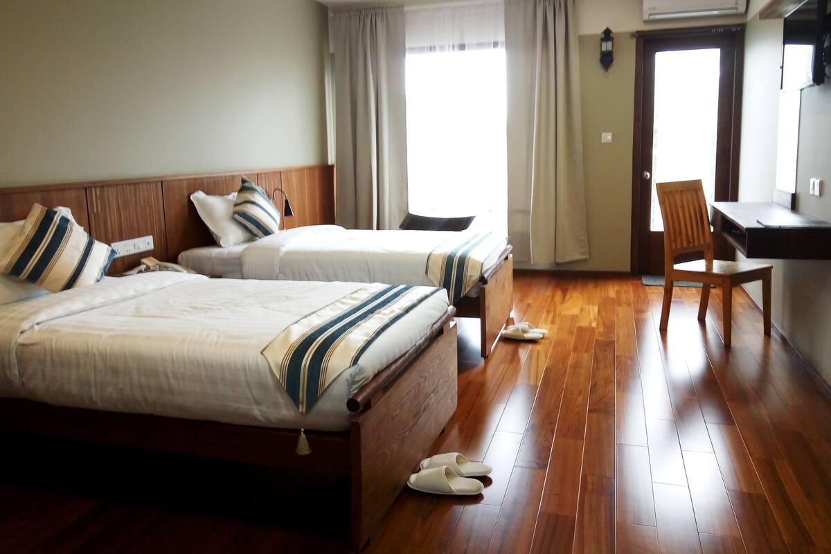A hotel room with two beds, a wooden floor, a desk, a chair, and a window with curtains.