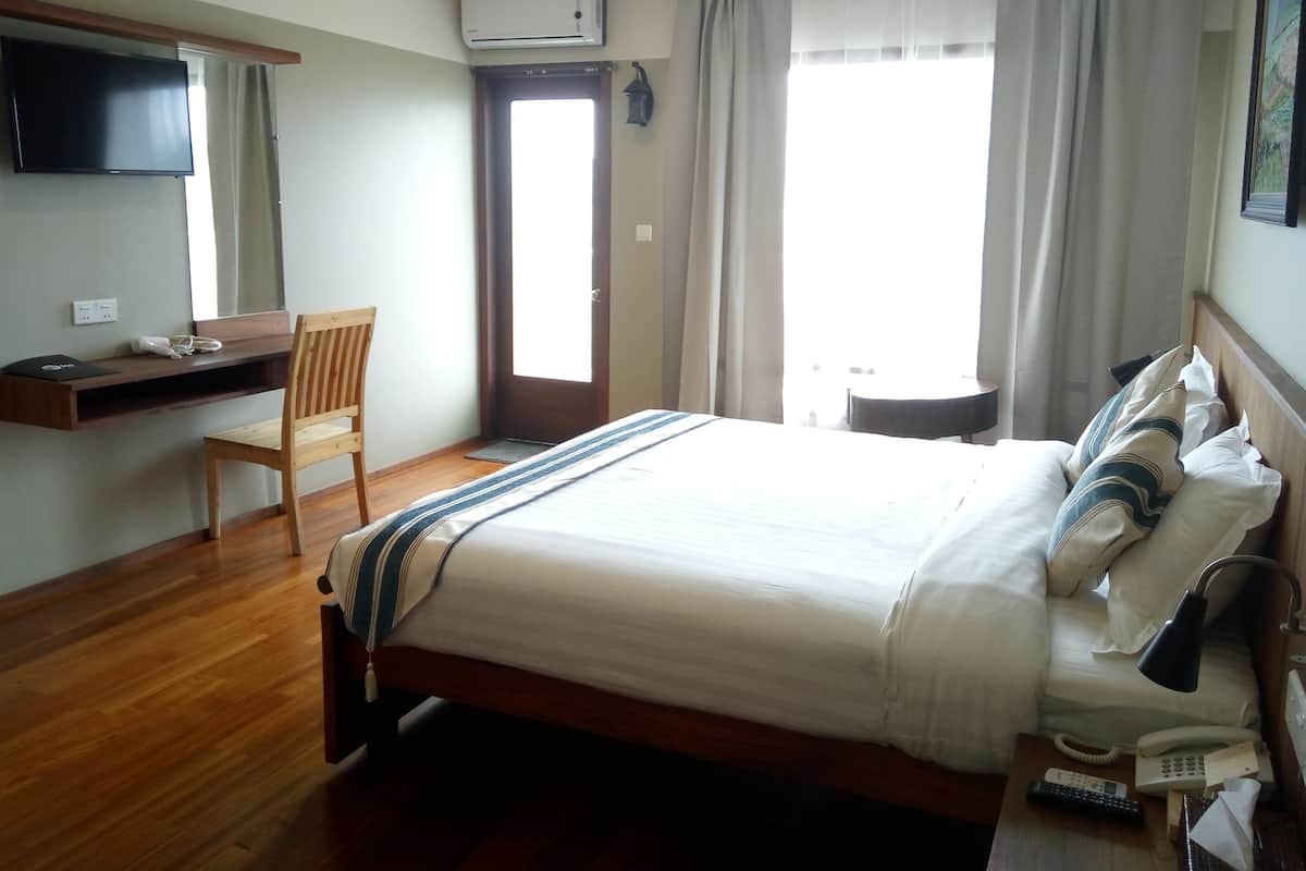 A hotel room with a bed, a desk with a chair, a TV, an air conditioner, a window with curtains, and a bedside table with a lamp and a phone.