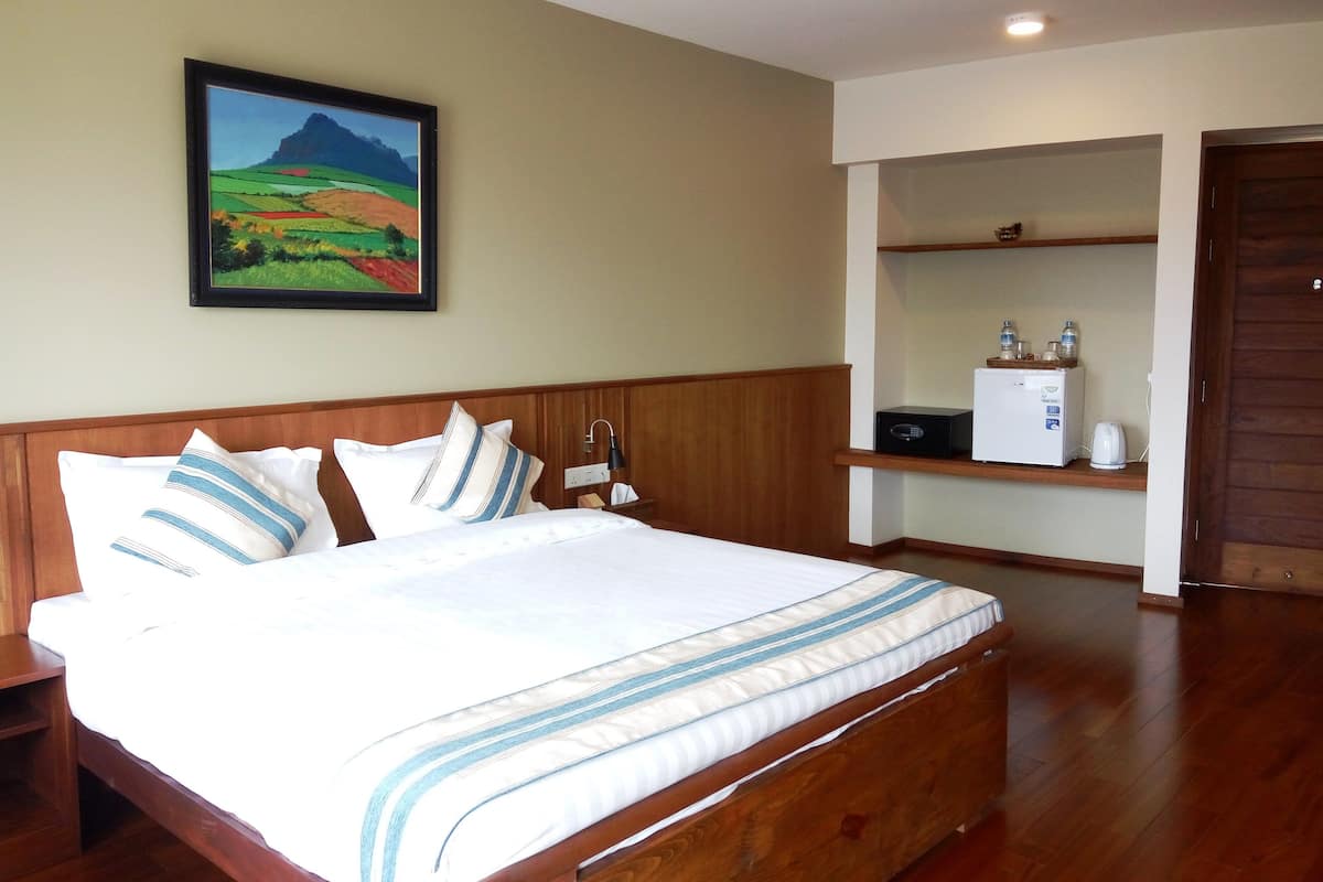 A hotel room with a wooden bed, a nightstand, a framed landscape painting, a shelf with a refrigerator and a kettle, and a wooden door.
