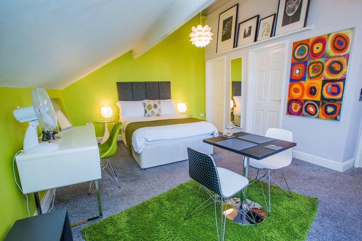 A modern hotel room with a bed, a desk, a chair, a small table, a fan, and a green rug.